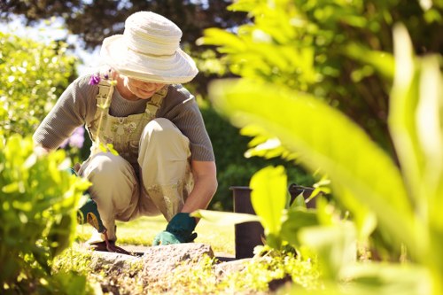 A woman in a garden during daylight hours is crouched down near a low stone border, tending to the soil and plants. She is wearing a wide-brimmed hat for sun protection, a light-colored apron, and gardening gloves. The garden features a well-maintained lawn with lush, green grass in the foreground, bordered by a neat stone edging separating it from flower beds and shrubbery. Behind her, there are various bushes and trees, with leaves in bright green and darker shades, indicating a healthy, cultivated outdoor space. Paved pathways and wooden decking are visible in the background, suggesting a landscaped yard typical of a residential property in Holborn or nearby areas. The scene is illuminated by natural sunlight, creating a warm and inviting atmosphere for outdoor gardening activities, which aligns with the gardening and landscaping services offered by Hedge Trimming Holborn.