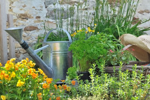 A vibrant garden scene featuring a variety of plants and garden elements, including a lush green bush with pink flowers, surrounded by smaller green foliage. In the foreground, there are bright yellow and orange flowering plants, adding lively colour to the garden. A silver metal watering can is placed near the plants, with its handle and spout visible, indicating ongoing garden maintenance. To the right, a straw hat rests on a wooden surface or garden furniture, suggesting a gardener taking a break. The background includes leafy green plants and tall grass-like vegetation, set against a light stone or brick wall that provides a rustic backdrop. The scene is well-lit, likely during a sunny day, highlighting the variety of textures—from the smooth watering can to the textured leaves and flowers. This outdoor garden space is part of a well-maintained yard, ideal for gardening and landscaping services such as hedge trimming and lawn care, as offered by Hedge Trimming Holborn, serving the Holborn area of London and nearby postcode regions.