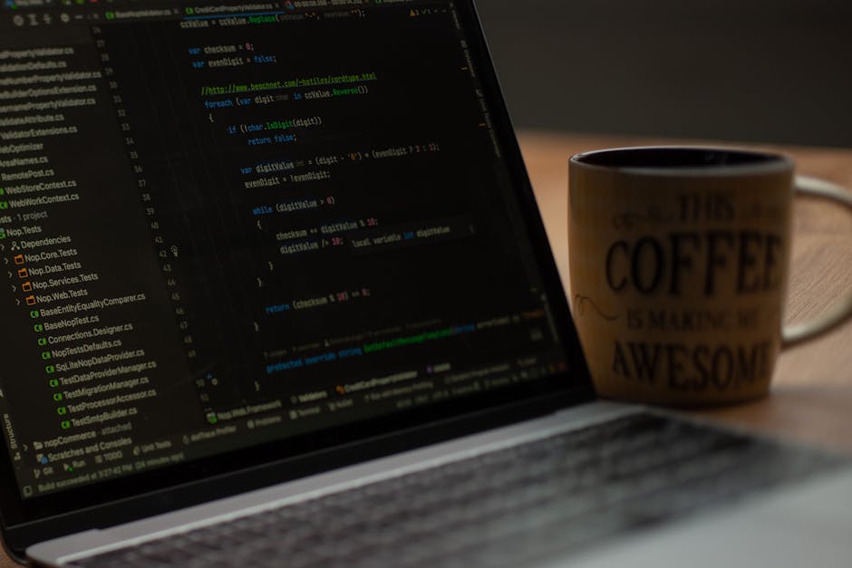 A close-up image of a laptop displaying lines of green and blue code on a dark background, placed on a wooden desk. To the right of the laptop, there is a ceramic mug with a beige background featuring