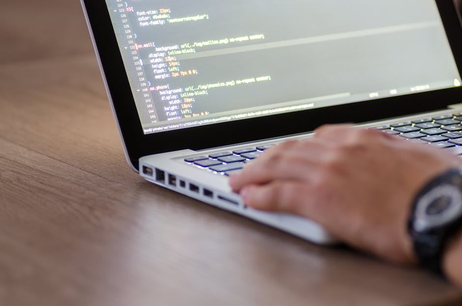 A close-up image of a laptop displaying lines of green and blue code on a dark background, placed on a wooden desk. To the right of the laptop, there is a ceramic mug with a beige background featuring